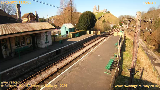 A view of a train station with a platform featuring several green benches. The station building is made of stone with a sloped roof and a chimney. In the background, a castle ruins are situated on a hill surrounded by trees. The sky is clear and blue, and there is a wooden shed on the platform. The railway tracks run parallel to the platform, leading into the distance. A wooden pole with electrical lines stands to the right of the image.