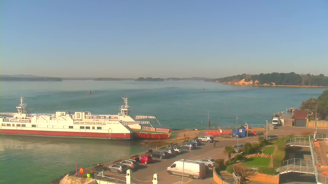 A ferry boat is docked at a harbor with a clear blue sky above. The water is calm and reflects the light, while a variety of vehicles are parked along the waterfront. There are trees and a sandy shore in the background, and a few distant islands are visible on the horizon. A small blue building and other structures are situated near the dock.