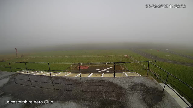 A foggy scene is depicted from a webcam at the Leicestershire Aero Club. The image shows a grassy airfield with a faint outline of the runway visible. In the foreground, there is a railing and a helipad marked with a white cross. A windsock on the left indicates low visibility due to the dense fog that obscures much of the background. The date and time are displayed at the top right corner.