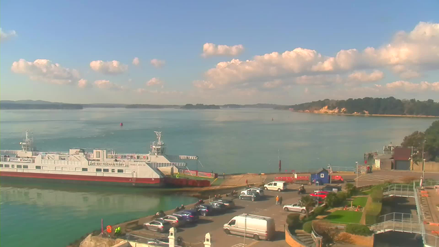 A serene coastal scene featuring a large ferry docked at a harbor. The ferry, painted white with red accents, is positioned near a parking lot filled with cars. In the background, calm blue water reflects fluffy white clouds in a clear sky. There are green hills and cliffs in the distance, and people can be seen walking along the waterfront. The shore is lined with greenery and pathways that lead down to the water.