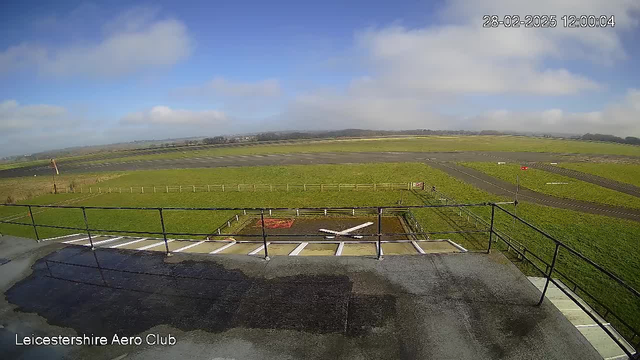 A view of a grassy airfield with a blue sky partly covered by clouds. In the foreground, there is a railing view from a platform, showing a white aircraft outline marked on the ground. The airfield extends into the distance, with several taxiways visible, bordered by wooden fencing and a few red flags. The date and time stamp at the top right shows 28-02-2025, 12:00:04.