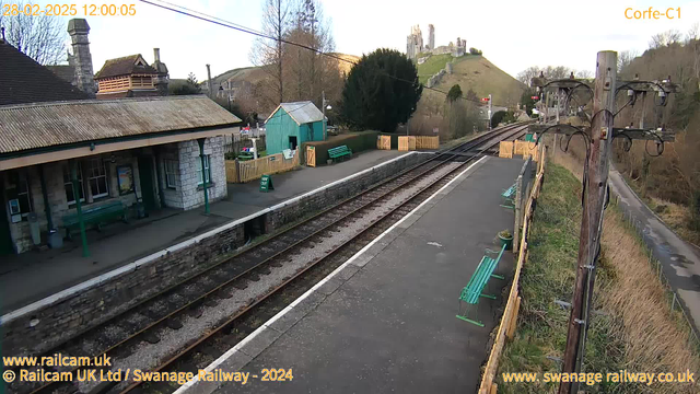 A railway station scene with tracks running through the center. On the left, there is a stone building with a peaked roof, supported by green-painted wooden pillars. A green bench is visible on the platform. Further along the right, there are more green benches and a wooden fence. In the background, a hill with ruins of a castle can be seen above the station. The sky is clear with a few clouds. A wooden post with electrical lines stands on the right side of the image.