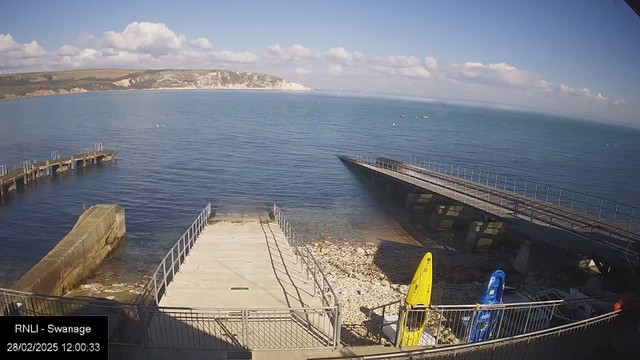 A coastal scene featuring calm blue waters and a cloudy sky. In the foreground, there is a stone jetty leading to the water, with a ramp on the right. Two kayaks, one yellow and one blue, are positioned on the shore next to the jetty. Small boats can be seen on the water in the distance, and the white cliffs are visible along the coastline. The setting is bright and sunny, suggesting a pleasant day by the sea.