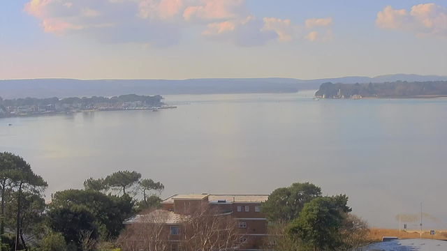 A serene waterfront view featuring calm waters reflecting soft pink and blue clouds in the sky. In the foreground, there are trees and a building with a flat roof. In the distance, several boats are docked along the shore, with a landscape of low hills and more trees lining the opposite bank of the water. The scene conveys a tranquil setting on a clear day.