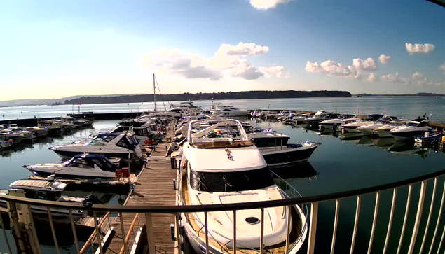 A tranquil marina filled with numerous boats docked at a wooden pier. The scene is illuminated by bright sunlight, casting reflections on the calm water. White fluffy clouds are scattered across a blue sky, and a distant shoreline is visible in the background. Some boats are lined up along the dock, while others are moored in the water.