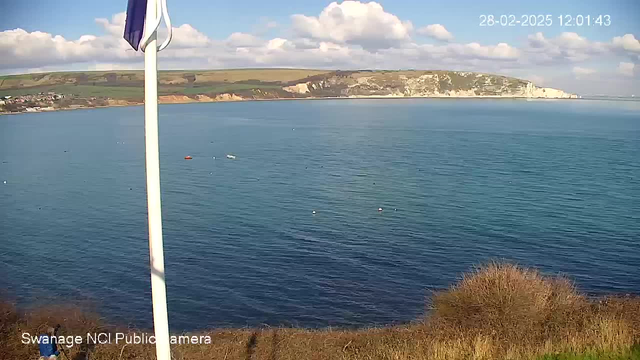 A coastal scene captured from a webcam shows calm blue waters under a clear sky with scattered white clouds. In the foreground, there is a flagpole with a flag. The shoreline features green hills, with a rocky cliff on the right. A few small boats are visible in the water, along with floating buoys. The date and time at the top right corner indicate it's February 28, 2025, at 12:01:43.