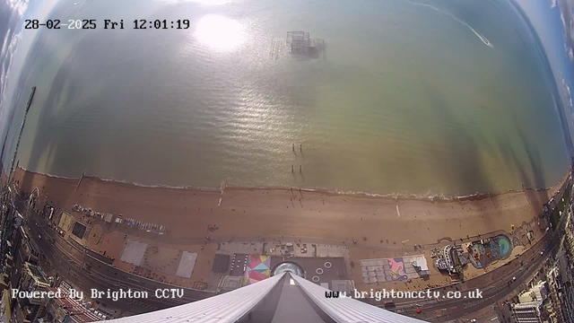 A high-angle view of a sandy beach with a few people visible along the shore. The ocean stretches out to the horizon, reflecting sunlight. In the distance, there is a pier with a structure partially submerged in the water. To the left, there are colorful attractions and buildings along the waterfront, while the lower part of the image features the rooftop edge and architectural details of a tall structure. The image is timestamped showing it is midday on February 28, 2025.