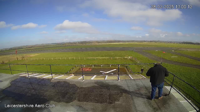 A view from a rooftop overlooking a grassy airfield with a runway. In the foreground, a person wearing a black jacket stands near a railing, looking out at the landscape. There is a white cross on the ground, possibly indicating a landing zone. In the background, there are clouds in a blue sky, and a small road with a fence to the left. The airfield is mostly green grass with some dark patches and a few buildings visible in the distance.