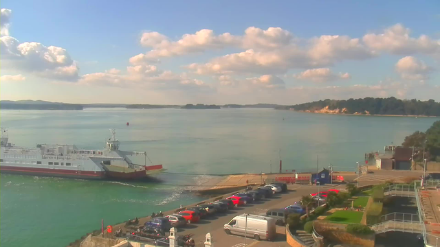 A ferry is approaching a dock, with its ramp lowered toward the pier. The water is calm, reflecting the blue sky above, which has fluffy white clouds. In the foreground, there is a parking area filled with cars of various colors, including red and silver. To the right, greenery and a landscaped area are visible, while a building with a dark roof is near the water's edge. The landscape includes distant wooded hills and a few vessels on the water.