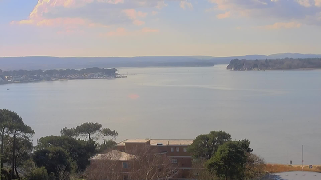 A tranquil landscape featuring a wide, calm water body reflecting soft sunlight, surrounded by distant hills. In the foreground, there are trees and a building with a flat roof, partially obscured by foliage. The shoreline in the background shows a marina with boats and a cluster of calm, rolling hills beyond the water, under a sky with soft clouds and a hint of blue.