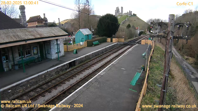 A view of a train station with stone walls and a covered waiting area. In the foreground, there are train tracks leading to the left and right, with green benches lining the platform. To the left, there’s a small green shed and a sign that reads "WAY OUT." In the background, a large hill is visible, topped with a historical castle ruins. Trees and shrubs surround the area, with clear skies above. The image is timestamped at 1:00 PM on February 28, 2025.