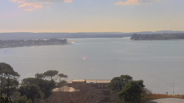 A panoramic view of a calm body of water reflecting the soft hues of a sunset. In the distance, a shoreline shows a mix of trees and buildings, with small boats scattered across the water. Colorful light reflections can be seen on the surface, and gentle hills rise in the background under a light blue sky with scattered clouds.