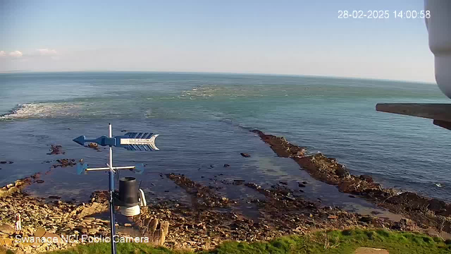 A scenic coastal view featuring a rocky shoreline. In the foreground, there is a weather vane with a blue arrow pointing left and a circular base. The background shows clear blue water stretching to the horizon, with a few scattered rocks and gentle waves. The sky is bright, with no clouds visible. A small figure is standing on the rocks near the water. The date and time are indicated in the upper right corner.