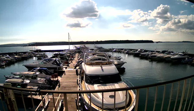 A marina filled with numerous boats docked along a wooden pier. Most boats are motorboats of varying sizes, with some displaying colorful covers. The water is calm and reflects the blue sky, which is scattered with fluffy white clouds. In the background, there are green hills and a distant shoreline, creating a peaceful waterfront scene.