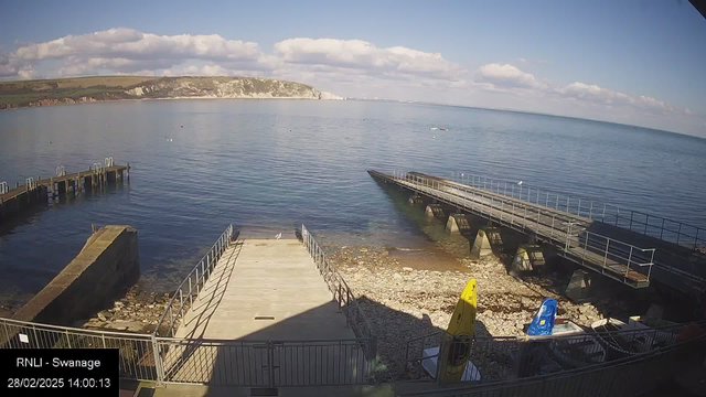 A view of a calm sea with gentle waves under a clear blue sky, dotted with a few fluffy clouds. In the foreground, there is a wooden jetty leading out over the water, with a stone walkway beside it. To the right, there are two small boats, one yellow and one blue, secured on dry land near the water's edge. The rocky shoreline is visible, and the landscape in the distance features green hills and white chalk cliffs.