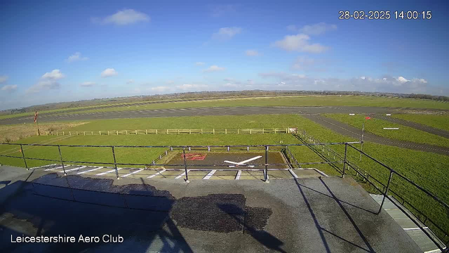 A clear blue sky with a few white clouds is visible above a wide green field. In the foreground, there is a black railing and a section of the roof from which the image is taken. Below, the ground includes a patch with a red "X" marking and small white lines. In the distance, a runway with a marked path is seen, bordered by a wooden fence. The scene is bright and well-lit, suggesting a sunny day.