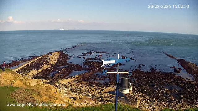 A coastal view featuring a rocky shoreline stretching into the calm sea. The sky is clear with some clouds. In the foreground, there is a weather vane pointing north with a small wind gauge attached to it. A person stands on the rocky edge near the water, while the grassy area is visible on the left side. The scene is well-lit, indicating a sunny day.