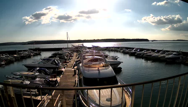 A marina scene featuring multiple boats docked along a wooden pier. Sunlight reflects off the water, creating a shimmering effect. In the background, a calm body of water extends towards a distant tree-lined shore under a partly cloudy sky. The foreground shows a sleek white boat with a cabin, among several other vessels of various sizes and colors.