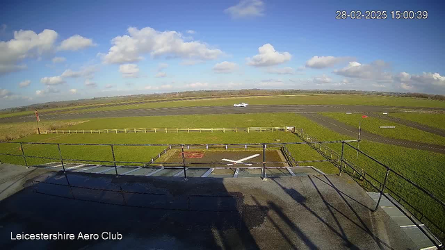 A wide view of an airfield under a clear blue sky with scattered clouds. In the foreground, there is a railing surrounding a helipad marked with a white cross, with a grey surface. Beyond the helipad, a grassy area stretches out, bordered by a white wooden fence. In the distance, there is a runway where a small white aircraft is taking off. The background includes expansive green fields and patches of trees. The image displays the timestamp "28-02-2025 15:00:39" in the corner.