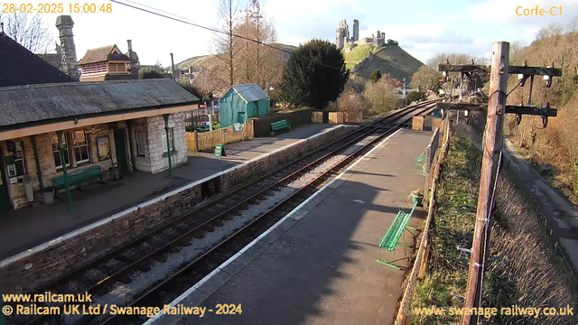 A railway station scene captured from a webcam, showing a platform with a stone and brick building on the left, featuring a sloped roof and several windows. There are green benches along the platform, and a wooden fence is visible in the background. To the right, railway tracks extend into the distance. In the background, a green hill rises, topped by the ruins of a castle. Trees and bushes are scattered throughout the area. The image is set during the day with clear skies.