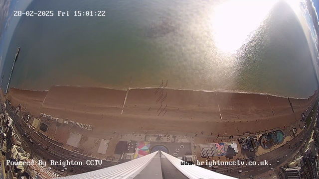 A high-angle view from a webcam overlooking the coastline of Brighton. The image captures a sandy beach with a gentle wave lapping at the shore, reflecting sunlight. There are several people walking along the beach, with distinct footprints visible in the sand. To the left, a pier extends into the sea, while colorful beach huts and an amusement area can be seen below. The sky is clear with a few clouds, and the time and date are displayed in the top left corner.