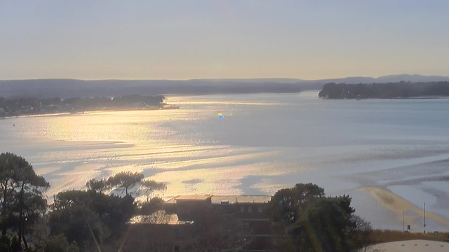 A tranquil water scene during the day, showing a shimmering surface reflecting sunlight. The background features light hills and trees along the shoreline, with some buildings visible in the foreground. A small, colorful reflection can be seen on the water, suggesting some activity or object in the distance. The sky is clear with a soft light indicating morning or late afternoon hours.