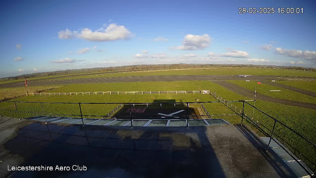 A wide view of an airfield from a high vantage point. In the foreground, there is a helipad marked with a white cross and surrounded by a fence. Beyond the helipad, a green grass field extends towards the horizon, with a parked small aircraft visible to the right on the runway. The sky is mostly clear with scattered white clouds, and the scene is bright, indicating it is daytime. In the bottom left corner, the text "Leicestershire Aero Club" is displayed.