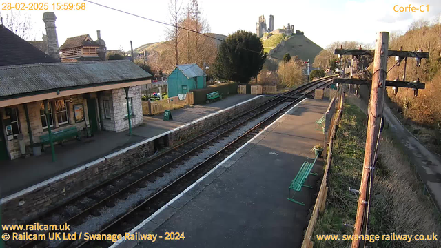 A view of a railway station set in a rural area. The foreground features a platform with two green benches, and a stone building, likely the station house, with a sloped roof. On the left, there is a small green shed and a picket fence. In the background, a hill rises sharply, topped by stone ruins, possibly a castle. The sky is clear with soft lighting, suggesting late afternoon. The railway tracks extend into the distance, surrounded by grassy areas and sparse trees.