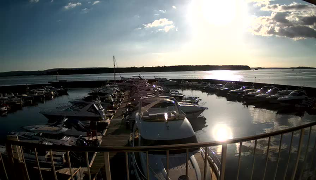 A marina with several boats docked in calm water, reflecting the bright sunlight. In the background, a shoreline with trees and a clear sky is visible, while a few fluffy clouds drift by. The scene conveys a serene, sunny day by the water.