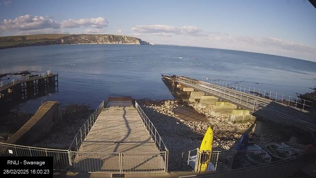 A scenic view of a calm seaside with clear blue water reflecting the sky. In the foreground, there are two piers made of wood, one leading into the water and the other slightly submerged. The shoreline is rocky with some pebbles and small boats visible on the left side. There are a few boats in the water and grassy hills in the background, with cliffs rising up on the horizon under a partly cloudy sky. The time and date are displayed at the bottom left.