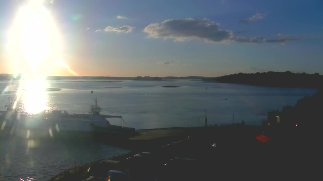 A serene waterfront scene during sunset, with the sun casting a bright reflection on the water. A ferry is docked at the shore on the left side of the image. The background features small islands and gentle hills under a clear sky scattered with a few clouds. The shoreline has some parked vehicles, and the setting sun creates a warm glow over the landscape, enhancing the tranquil atmosphere of the scene.