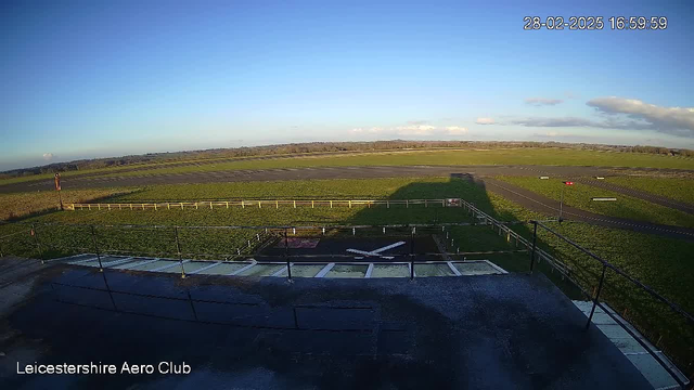 A wide view of an open field and runway at Leicestershire Aero Club during daylight. The sky is clear with a few clouds. The foreground features a tarmac area with markings, including a large white cross. In the distance, there is a boundary fence and grassy areas, while the runway is visible in the background. The image is time-stamped with the date and time at the top right corner.