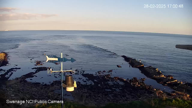 A calm ocean scene under a clear sky, with gentle waves lapping at rocky shores. In the foreground, there is a weather vane on a post, indicating wind direction. A few people are standing on a rocky outcrop near the water. The sun is low in the sky, casting soft lighting over the landscape.