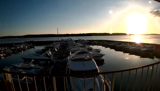 A marina at sunset, featuring numerous boats docked in still water. The sun is low on the horizon, creating a warm, glowing reflection on the water's surface. The sky transitions from blue to orange and yellow, with a few clouds scattered. Near the foreground, a railing frames the view. In the background, a shoreline with trees is silhouetted against the bright sky.