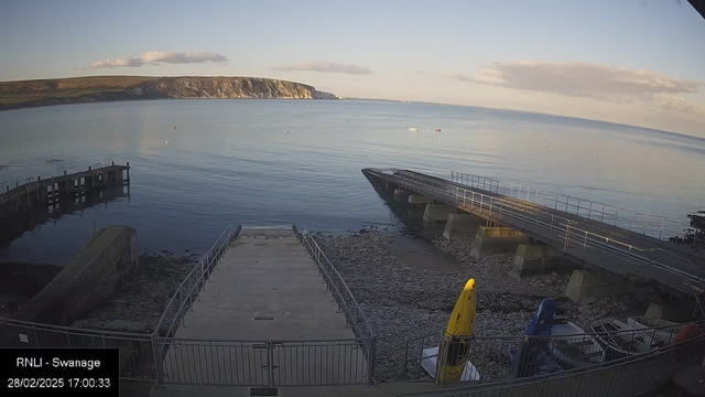 A calm seaside view at Swanage. The image shows two piers extending into the water, with one pier featuring a wide ramp leading down to a rocky shore. In the foreground, a bright yellow kayak is positioned on the right side, while a blue boat is partially visible next to it. The water is still with a few small boats in the distance, and the cliffs are visible in the background under a clear sky. The overall scene conveys a serene coastal atmosphere.