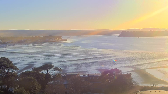 A sunrise over a calm body of water with soft waves reflects light. The scene includes distant hills and a shoreline dotted with trees. In the foreground, there are low-lying buildings surrounded by greenery. Soft pastel colors blend in the sky, suggesting early morning.