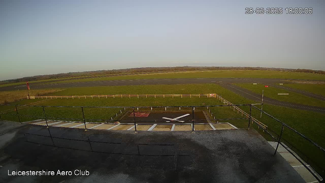 A wide view of a grassy airfield under a clear sky. In the foreground, there is a flat rooftop with a railing, and a white X marking on the ground, likely indicating a helipad or landing area. To the left, a structure resembles a small control tower or building. The airfield stretches out towards the horizon, with a runway visible, and a few scattered markers along the ground. The scene is tranquil, captured during sunset with soft lighting.