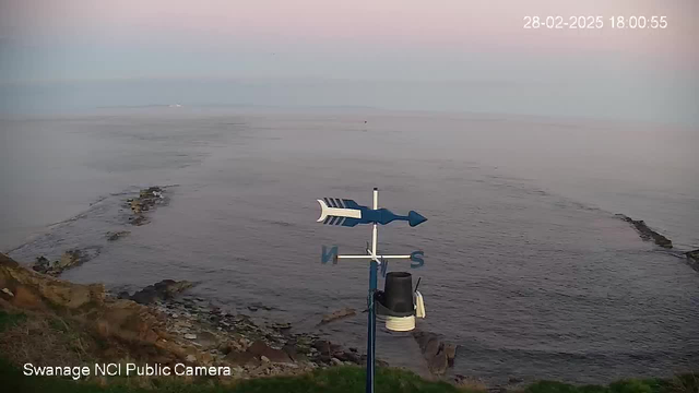 A coastal view showing a calm sea under a pastel sky. In the foreground, there is a blue weather vane with arrows pointing north and south. The shoreline contains scattered rocks and grass, with a few rocky outcrops extending into the water. The image captures a serene, twilight atmosphere.