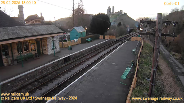 A view of Corfe Castle station during sunset. The image shows a train platform lined with green benches. On the left, there is a building with a sloped roof, featuring a sign that reads "Corfe Castle." A small blue building and a wooden fence are visible in the background, along with trees and the outline of Corfe Castle on a hilltop. The railway tracks run parallel to the platform, and the scene is quiet with a soft evening light.