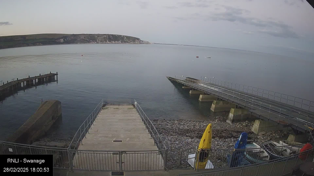 A calm seaside scene shows a wide expanse of water reflecting the sky, with gentle ripples on the surface. In the foreground, there is a concrete ramp leading down to the water, surrounded by a railing. To the left, a wooden jetty extends into the water, with a mix of solid and grid surfaces. On the right, several colorful boats are visible, including a yellow and a blue kayak. Pebbles and a small beach line the edge of the ramp. In the background, grassy hills and a distant cliff outline the shore under a partly cloudy sky.