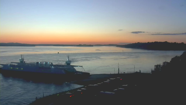 A serene evening scene featuring a calm body of water reflecting soft hues of blue, orange, and pink from the sunset. In the foreground, a large ferry boat is docked, illuminated by lights. Nearby, several parked cars are visible along the water's edge, with a faint shoreline and trees in the background silhouetted against the colorful sky. The atmosphere is tranquil, with gentle waves and hints of distant land across the water.