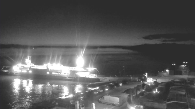 A dark scene showing a partially illuminated ferry docked at a pier. Bright lights on the ferry reflect on the water's surface, creating a shimmering effect. The surrounding area includes parked vehicles and some structures, with faint outlines of hills visible in the distance against the night sky. The atmosphere is calm and serene, with a hint of fog over the water.