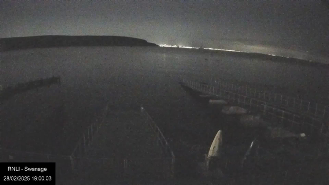 A dimly lit scene of a calm body of water at night. In the foreground, there is a wooden structure or dock extending into the water, with some boats moored alongside it. The background features a distant shoreline illuminated by faint lights, suggesting a town or city. The sky is mostly dark with a hint of light on the horizon. The timestamp indicates the date and time of the image.