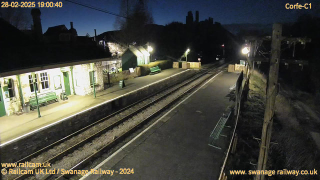 A dimly lit train station platform at dusk, featuring two sets of railway tracks converging towards the right. On the left, there is a stone building with a green roof and wooden benches outside. A sign reading "WAY OUT" is seen near a wooden fence. There are several benches along the platform, and trees can be seen in the background. The sky is darkening, and a few lights illuminate the station.