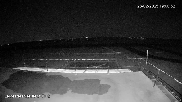 A dark scene captured from a webcam at the Leicestershire Aero Club shows a flat surface in the foreground with a large rectangular area marked out. The surface has a wet patch and two lines of light-colored sections, which may represent hangars or buildings. In the background, faint lights from distant buildings or vehicles can be seen against the dark sky. The date and time indicate it is evening.