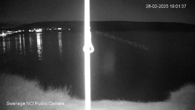 A dark image captured by a webcam shows a body of water reflecting faint lights from the distant shoreline. On the left side, a white pole is visible, standing against the night sky. The scene conveys a calm and quiet atmosphere with minimal visible activity. The time displayed in the top right corner indicates it is shortly after 7 PM.