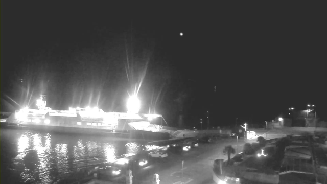 A brightly lit ferry is docked at a port under a dark sky. The ferry has several bright lights illuminating its deck and the surrounding water, which reflects the lights. In the foreground, various vehicles are parked along the waterfront, with additional lights visible on the shore, indicating nearby structures. The image conveys a nighttime scene with strong contrasts between the light from the ferry and the darkness of the surroundings.