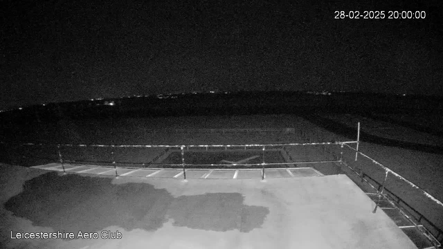 A dark scene captured from a webcam at the Leicestershire Aero Club. The foreground shows a flat surface with some indistinct shapes, possibly water, on the left side. A railing is visible in the middle of the image, extending horizontally across the scene. In the background, there are faint lights in the distance against an otherwise dark sky. The image is timestamped "28-02-2025 20:00:00."