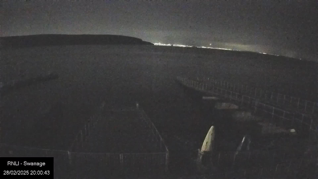 A dimly lit coastal scene at night, featuring calm water extending toward a dark horizon. In the foreground, there are moored boats or kayaks, partially visible on a dock. The background contains a distant shoreline illuminated by faint lights, suggesting a nearby town or city. The atmosphere is tranquil, with clouds obscuring the moonlight. A timestamp is visible in the lower left corner indicating the date and time.