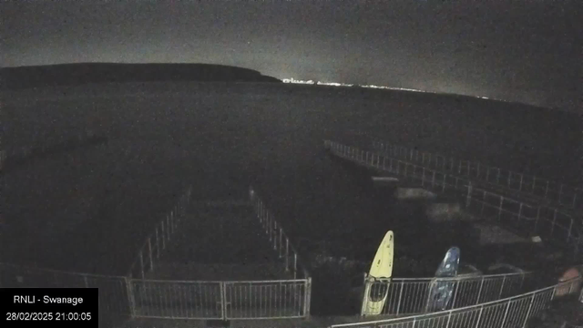 The image shows a dark sky above a calm body of water, with a distant landmass visible on the horizon. In the foreground, there is a fenced area with two kayaks resting on the ground. One kayak is yellow and the other is blue. A pier extends into the water but is barely visible due to the low light conditions. The overall scene appears quiet and still, suggesting nighttime.